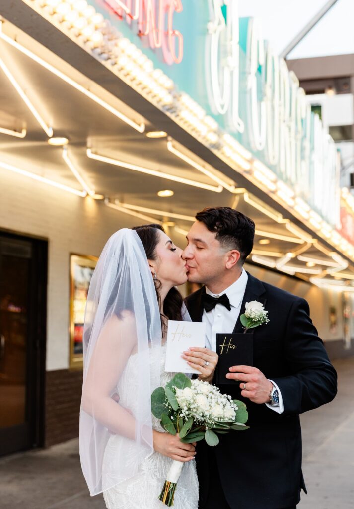 Newlyweds kissing at Fremont Street after Vegas ceremony while holding their vows booklet